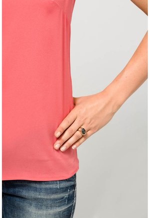 Main droite avec une bague en or et une pierre précieuse noire reposant sur la hanche, portant une chemise rose et un jean bleu sur un fond clair uni.