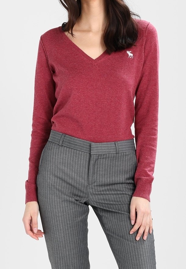 Femme portant un pull ajusté rouge à col en V et un pantalon gris à fines rayures, debout avec une main détendue le long du corps.
