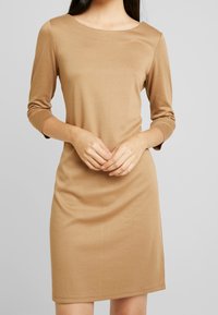 Abito marrone, aderente, con scollatura tonda e maniche a tre quarti, realizzato in un materiale liscio e elastico. Finisce sopra il ginocchio.