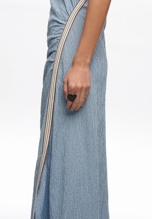 Widok z boku kobiety w jasnoniebieskiej plisowanej spódnicy z beżowym paskiem, pokazującej dłoń z dużym czarnym pierścieniem w kształcie serca.