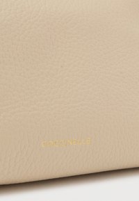 Sac à main en cuir beige avec une surface texturée et une marque en or. Vue rapprochée mettant en valeur le matériau doux et le motif subtil.