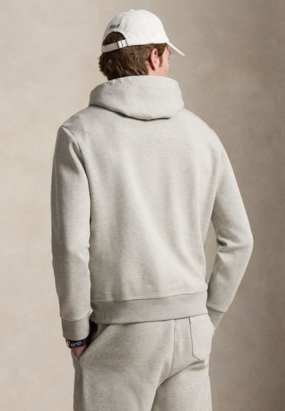 Homme portant un sweat à capuche gris clair et un pantalon de survêtement, casquette Polo blanche et montre-bracelet sombre, debout les mains dans les poches, dos tourné.