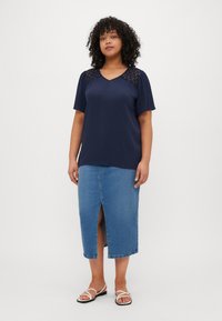 Navy blouse with lace detail on shoulders, v-neckline; paired with a light blue denim skirt featuring a front slit, and white sandals.