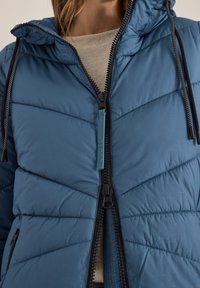 Giacca blu imbottita con cappuccio, dotata di una cerniera a tutta lunghezza e dettagli neri. Tessuto texturizzato con linee di cucitura diagonali.