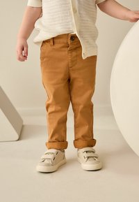 Pantalons en coton marron avec des ourlets retroussés, associés à des baskets beige à velcro. Le haut est composé d'un cardigan tricoté blanc avec des boutons.