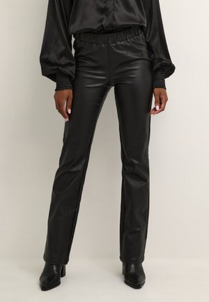 Person trägt schwarze, gerade geschnittene Hose aus Kunstleder, schwarze Langarmbluse und schwarze spitz zulaufende Stiefeletten vor einfarbigem Hintergrund.