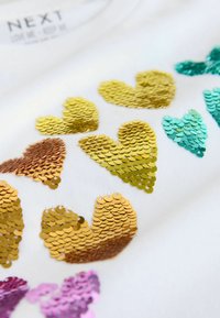 Tela blanca con corazones multicolores de lentejuelas en dorado, turquesa y rosa. Las lentejuelas son brillantes y de tamaños variados, creando textura.