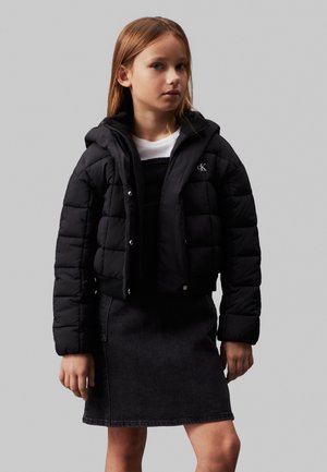 Giubbotto imbottito nero con chiusura a zip e a scatto, dotato di cappuccio, taglio ampio e logo. Abbinato a una gonna di jeans scuri e a una maglietta bianca.