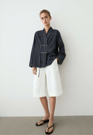 Jeune femme debout contre un mur blanc, portant une veste portefeuille en denim foncé, un short blanc à jambes larges et des sandales noires, mains détendues.