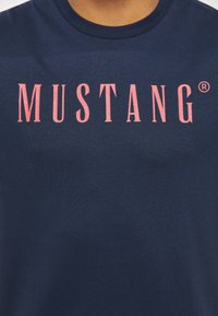 Navy blue cotton t-shirt featuring a large pink serif logo "MUSTANG®" on the front. The fabric appears smooth with a classic crew neckline.