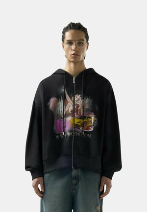 Uomo con capelli intrecciati e occhiali che indossa una felpa nera con zip con grafica di una donna dei cartoni animati, un'auto gialla, scatole rosa e testo.