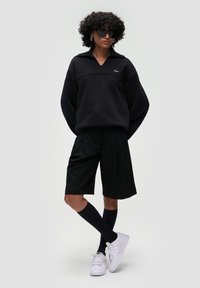 Felpa nera oversize con colletto, abbinata a pantaloni corti plissettati neri. Il modello indossa scarpe da ginnastica bianche e calze nere fino al ginocchio. Occhiali da sole neri.
