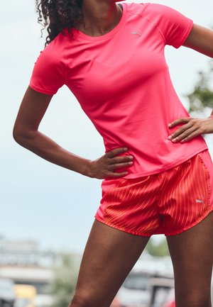 Woman in bright pink athletic shirt and matching shorts stretching outdoors with hands on hips against a blurred sky and trees background.