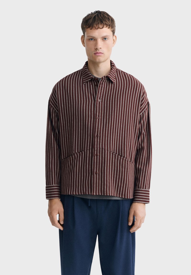 Jeune homme portant une chemise bordeaux à rayures verticales blanches et un pantalon bleu marine, debout devant un fond gris clair uni.