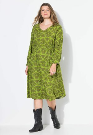 Grünes Kleid mit langen Ärmeln und V-Ausschnitt, mit floralem Muster und gerafft an der Taille. Kombiniert mit schwarzen Cowboy-Stiefeln.