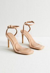 Nude högklackade sandaler med en klar, transparent rem. Fyrkantig tådesign, ankelspänne och smal stilettklack. Slät textur genomgående.