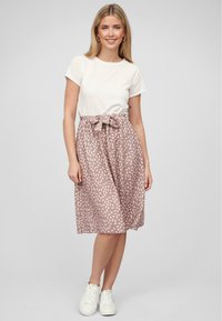 T-shirt blanc léger associé à une jupe rose pâle de longueur genou avec un motif de feuilles et un détail de taille à nouer. Des baskets blanches complètent le look.