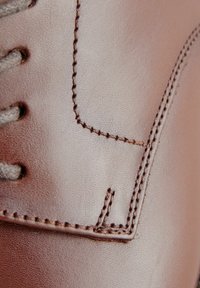 Gros plan sur une chaussure en cuir marron montrant une surface texturée, un contraste avec des coutures marron, et un œillet pour les lacets. Lisse avec une finition mate.