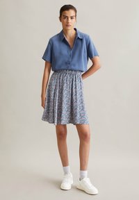 Blaues kurzärmliges Hemd mit Knöpfen, kombiniert mit einem floral bedruckten, gefältelten Rock. Weiße Sneakers und knöchellange Socken runden das Outfit ab.