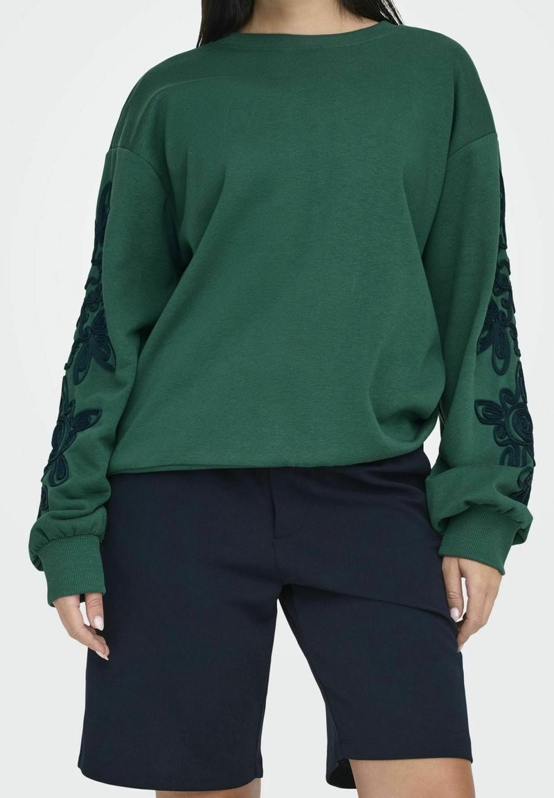 Sudadera de color verde oscuro de manga larga con patrones de flores bordados en las mangas, combinada con pantalones cortos de color azul marino oscuro.