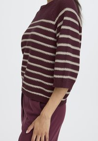 Burgundy and beige striped sweater with a round neck, short sleeves, and textured knit pattern, worn with tailored maroon trousers.