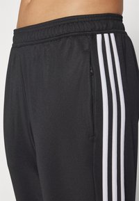 Zwarte sportbroek met een gestructureerde stof, elastische tailleband en drie witte verticale strepen langs de buitennaad. Met rits afgesloten zakdetails.