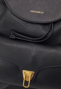 Zwarte leren tas met textuur, voorzien van een gouden logo, een metalen sluiting en een gebogen flapdetail, dat een hedendaags ontwerp benadrukt.