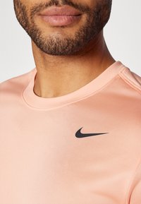Vaaleanpunainen Nike-urheilupaita, jossa on pyöreä pääntie ja musta logo vasemmalla puolella. Paita on valmistettu sileästä ja hengittävästä materiaalista.