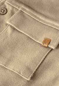 Kapsa na košili z béžové rypsové tkaniny s klopou, knoflíkem a malou hnědou koženou cedulkou přišitou na okraji.