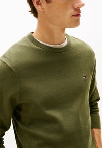 Maglione crew neck verde oliva realizzato in tessuto lavorato a maglia con una piccola patch del logo caratterizzata da accenti rossi, bianchi e blu sul petto.