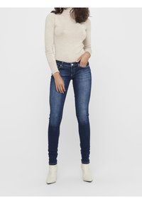 Úzké modré džíny vyrobené z denimu, s jemným zesvětlením, pětipocket designem, a v kombinaci s bílými kotníkovými botami na světlém pozadí.