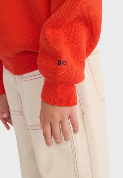 Personne portant un pull en maille orange avec "B-C" brodé sur le poignet de la manche et un pantalon clair avec des surpiqûres rouges.