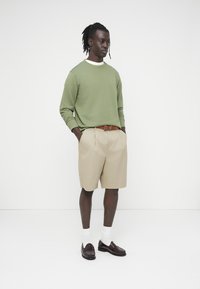 Ανοιχτό πράσινο μακρυμάνικο φούτερ, μπεζ σορτς με πιέτες, καφέ ζώνη, λευκές κάλτσες και σκοτεινά παπούτσια. Απλή, καθημερινή εμφάνιση με εφαρμοστή σχεδίαση.