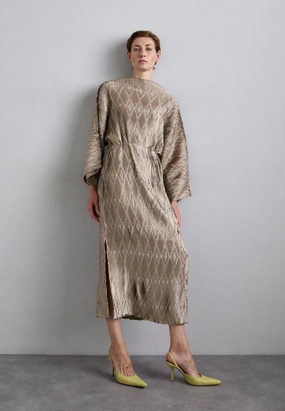 Woman standing against gray wall wearing textured beige midi dress with slit and yellow high-heeled slingback shoes.