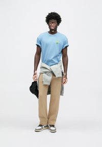 Gaiši zils īsroku T-krekls ar zaļu logotipu, pelēks džemperis sasiets ap vidukli, bēšas platām kājām bikses un pelēkas un melnas sporta kurpes.