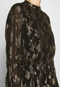 Robe noire avec broderie en feuilles métalliques dorées, col haut, tissu fluide et taille froncée, avec manches longues et texture transparente.