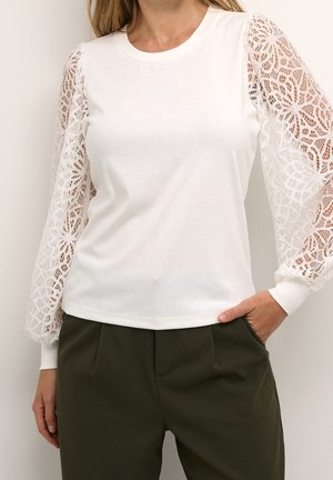 Femme portant un haut blanc à manches longues en dentelle florale et un pantalon taille haute vert olive, une main dans la poche.