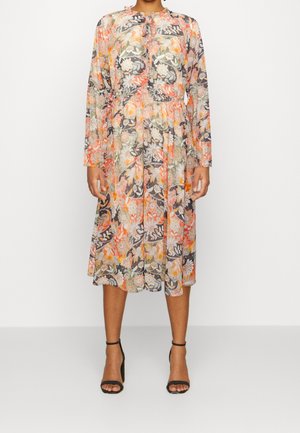 Frau trägt ein langärmliges, knielanges Blumenmusterkleid in Orange-, Schwarz-, Weiß- und Grüntönen mit schwarzen Knöchelriemen-Pumps.