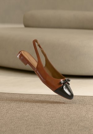 Bruine slingback platte schoen met zwarte neuskap en strikdetail, zwevend in de lucht boven een beige, gestructureerd tapijt met een neutrale bank op de achtergrond.