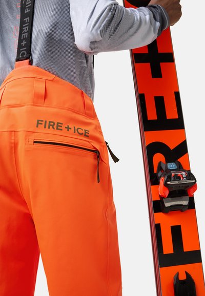 Jasnopomarańczowe spodnie narciarskie z regulowanymi czarnymi szelkami i tylną kieszenią na zamek, połączone z czarną i pomarańczową nartą. Trwa�ły, wodoodporny materiał.