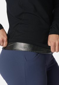 Person in einem schwarzen langärmligen Oberteil, das den Saum anhebt, um das reflektierende Netzfutter zu zeigen, kombiniert mit dunkelblauen, enganliegenden Hosen mit Taschen.