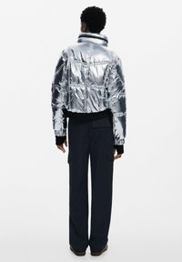 Giacca a vento argento con colletto alto, texture trapuntata e polsini neri, abbinata a pantaloni larghi blu navy e calzature scure.