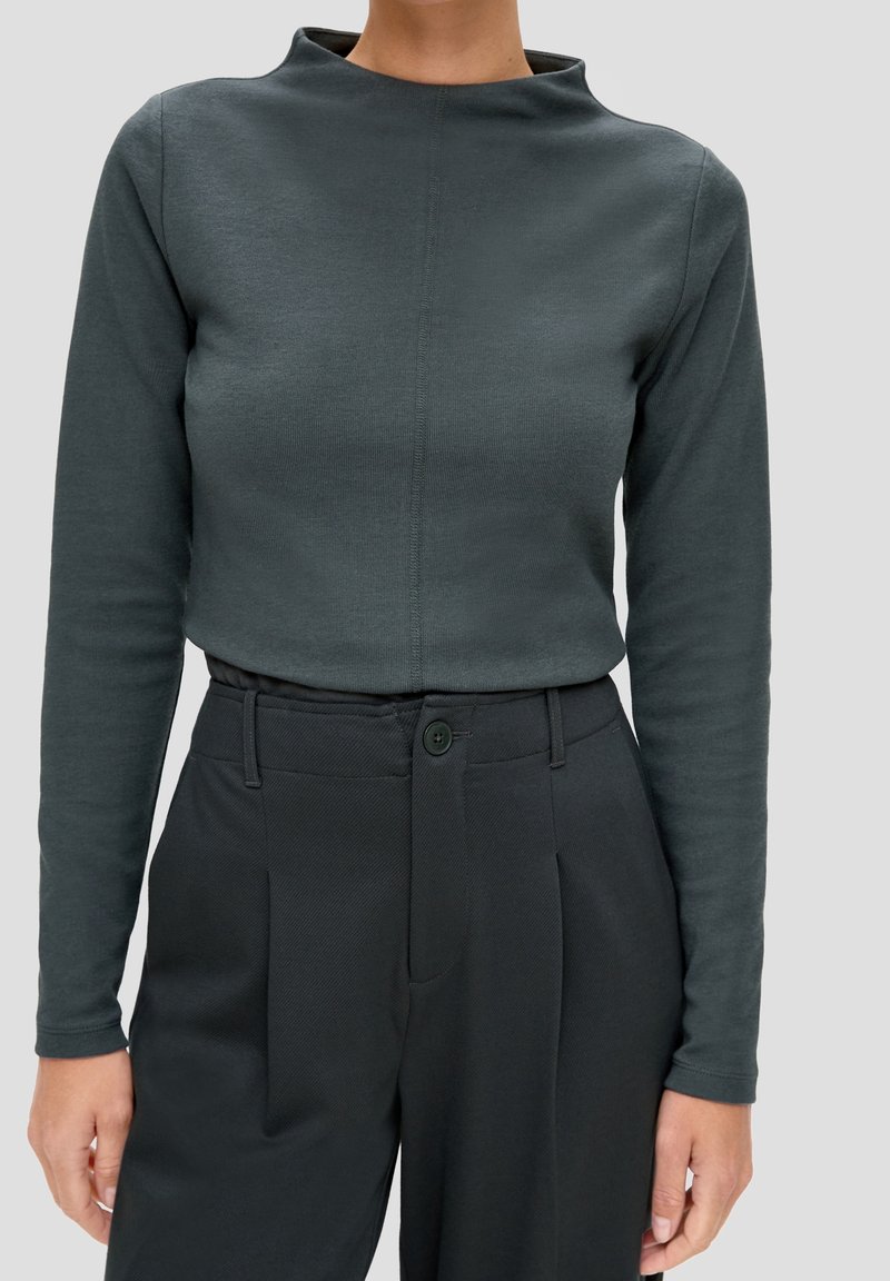 Maglietta a maniche lunghe di un verde scuro con un collo alto, caratterizzata da una texture liscia e da un design semplice, abbinata a pantaloni neri su misura.