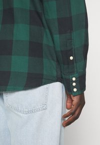 Zielona i czarna koszula w kratę z prostym dołem, zapinanym mankietem na trzy białe guziki oraz teksturowanym materiałem. Widoczne jasnoniebieskie dżinsy.