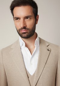 Mężczyzna z brodą i krótkimi ciemnymi włosami, ubrany w beżową marynarkę i białą koszulę, lekko spoglądający na bok na jednolitym tle.