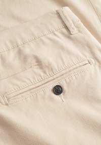 Beige stoffen broek met een achterzak met een horizontale opening, een zwarte knoop en gestikte details langs de randen.