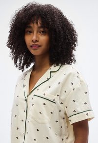 Κρεμ πουκάμισο με κοντά μανίκια και πράσινη επένδυση, με λουλουδένιο μοτίβο από μικρά κόκκινα και πράσινα σχέδια και με μπροστινή τσέπη.