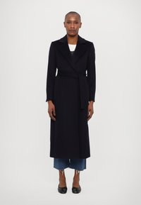 Dlouhý černý kabát z vlněné směsi s klopovým límcem, páskem v pase a plnými rukávy, nošený přes zkrácené modré džíny s třásněmi a černé boty.