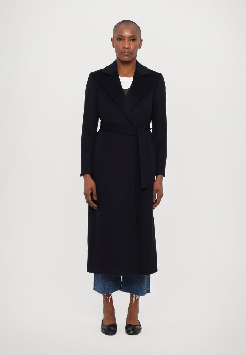 Dlouhý černý kabát z vlněné směsi s klopovým límcem, páskem v pase a plnými rukávy, nošený přes zkrácené modré džíny s třásněmi a černé boty.