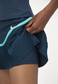 Falda deportiva azul marino con pliegues y una banda en la cintura de color turquesa como detalle. Al levantar los pliegues con la mano, se revela una capa interior lisa.